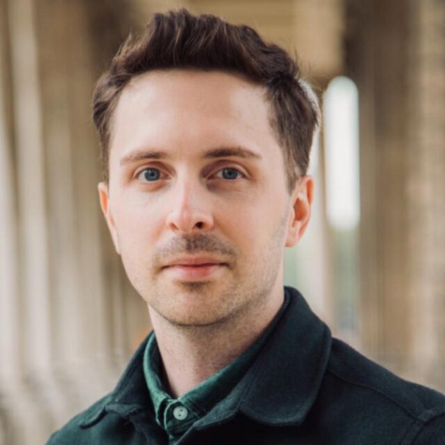 Man with short brown hair and blue eyes wearing a dark green jacket, standing in front of tall columns