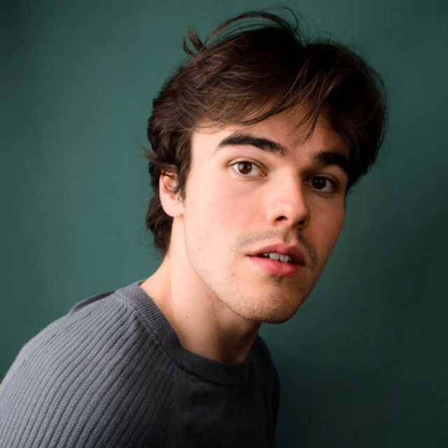 Young man with tousled brown hair in a grey jumper, looking over his shoulder against a teal backdrop