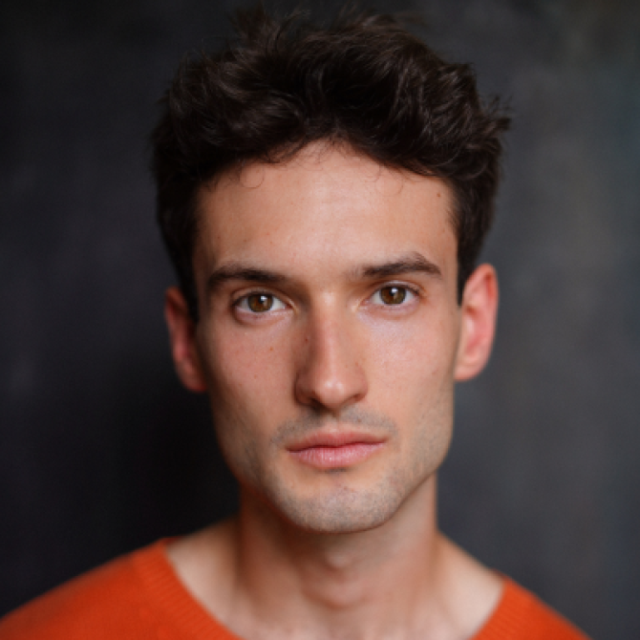 Portrait of a young man with dark curly hair, wearing an orange jumper, against a dark background