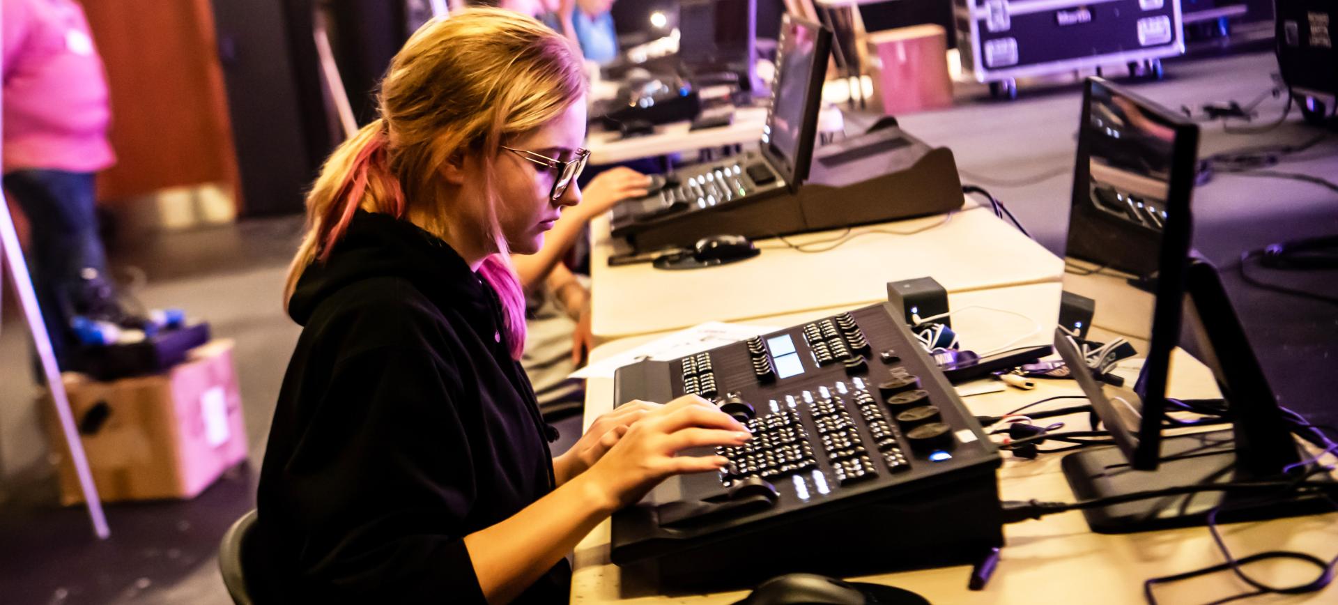 Person operating a lighting control console with sliders and buttons, monitors displaying show data nearby
