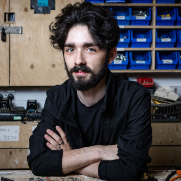 Man with curly hair and beard, arms crossed, sitting in a workshop with storage bins
