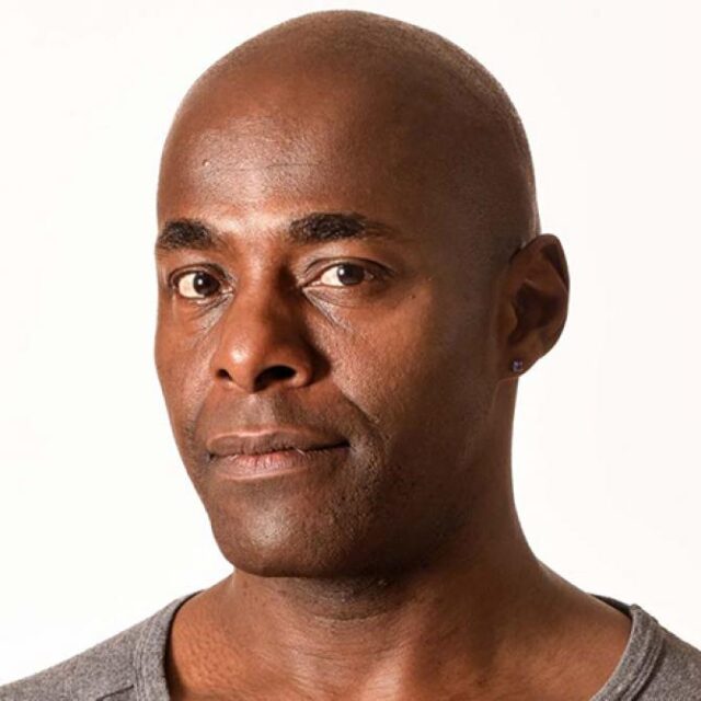 Headshot of a bald man with a moustache, wearing a grey top, against a plain light background
