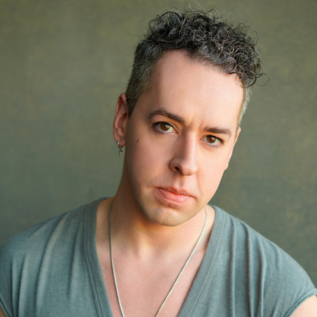 Portrait of a man with curly grey-flecked hair, wearing a green top and a beaded necklace, looking directly at the camera