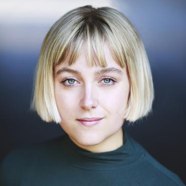 Headshot of a young woman with a blonde bob haircut and fringe, wearing a dark green top