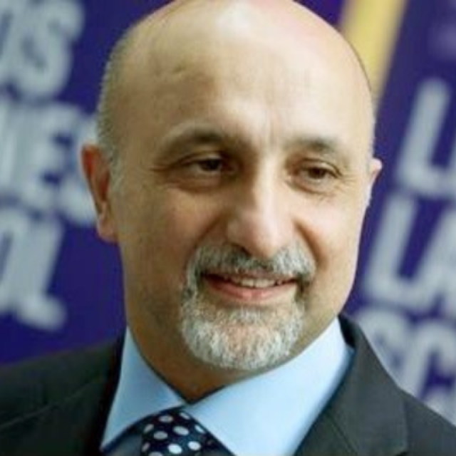 Bald man with a grey beard, smiling, wearing a dark suit and light blue shirt with patterned tie