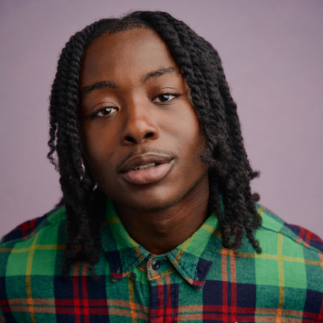 Young man with shoulder-length twisted locs wearing a green and red plaid shirt, looking at the camera