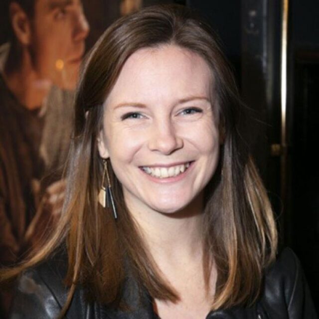 Woman with long brown hair smiling at a media event, wearing a black leather jacket