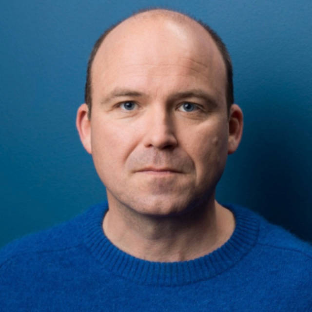 Headshot of a man with a shaved head wearing a blue jumper, against a teal background