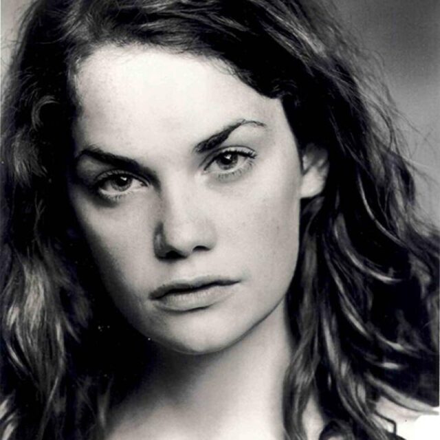 Black-and-white close-up portrait of a young woman with long curly hair, looking directly at the camera with a neutral expression