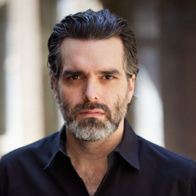 Man with greying hair and beard, wearing a dark shirt, looking directly at the camera with a serious expression
