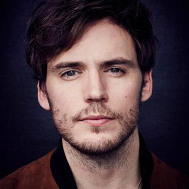 Close-up portrait of a young man with wavy dark hair and stubble, wearing a brown jacket against a dark background