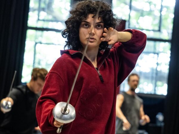 Woman in a maroon jumper holding a fencing foil in a training studio, with other fencers practising behind her