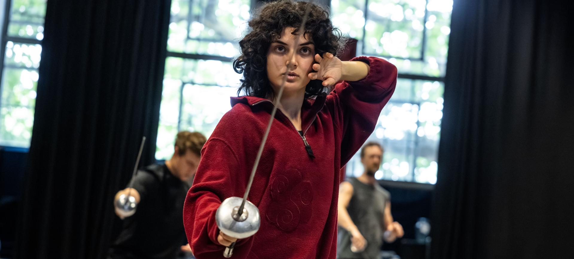 Woman in a maroon jumper holding a fencing foil in a training studio, with other fencers practising behind her