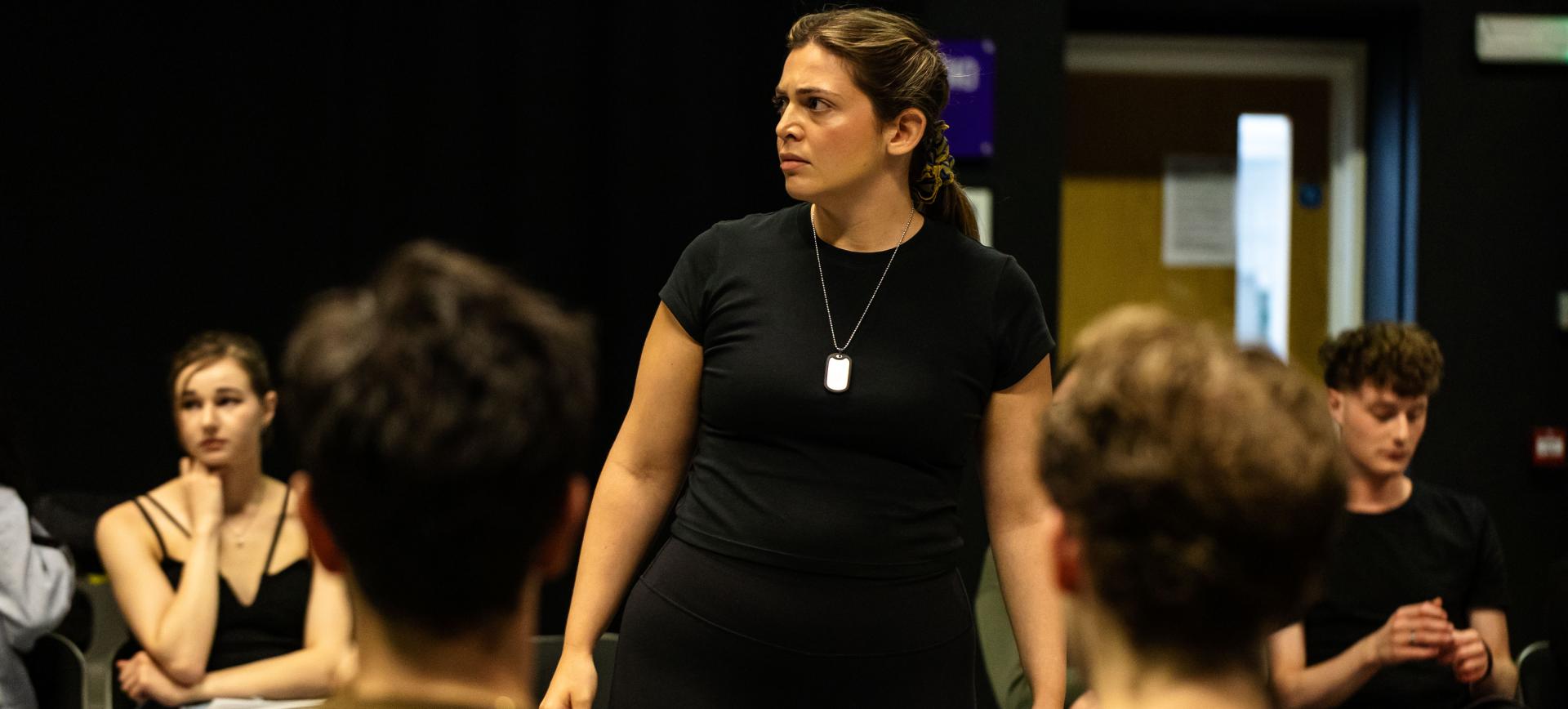 A woman stands addressing a seated group during a rehearsal or workshop session