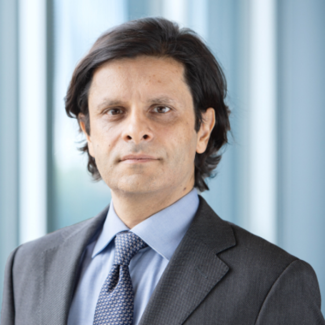 Portrait of a man with shoulder-length dark hair, wearing a grey suit and patterned tie