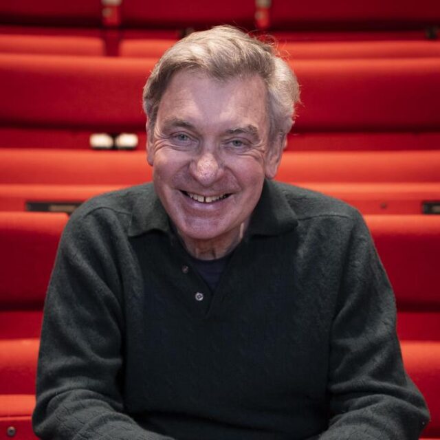 Older man with grey hair smiling warmly, seated in front of red theatre seating