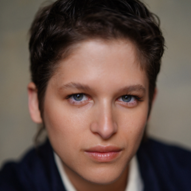 Close-up portrait of a person with short dark curly hair and blue eyes, looking directly at the camera