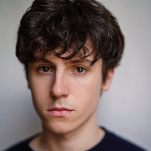 Close-up portrait of a young man with curly dark hair, looking directly at the camera with a neutral expression