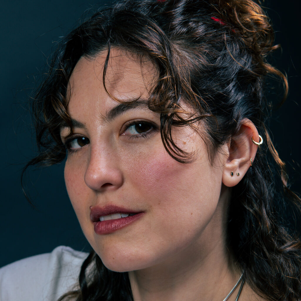 Close-up portrait of a woman with dark curly hair and multiple ear piercings, looking sideways at the camera