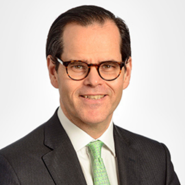 Portrait of a man with glasses, wearing a dark suit, white shirt and green patterned tie