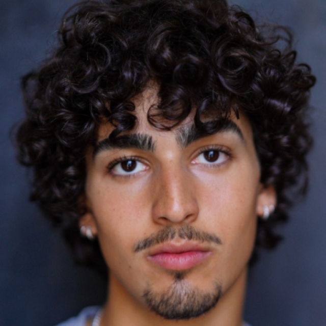 Close-up portrait of a young man with curly dark hair, light stubble and stud earrings, looking directly at the camera