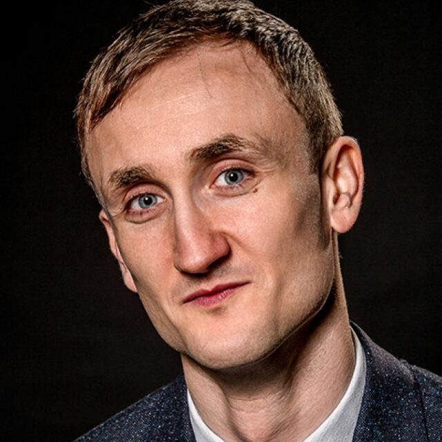 Portrait of a man with short brown hair, wearing a dark blazer over a white shirt, against a dark background