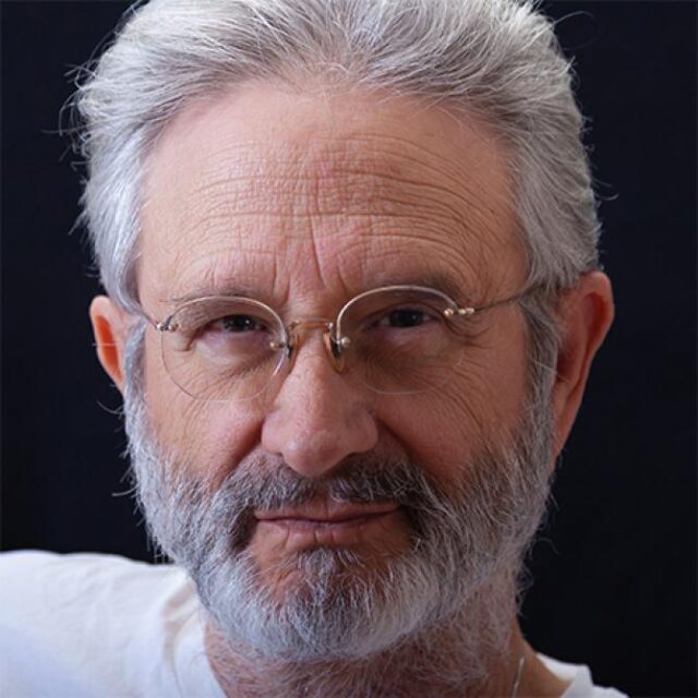 Close-up portrait of an older man with grey hair, beard and wire-rimmed glasses, wearing a white top