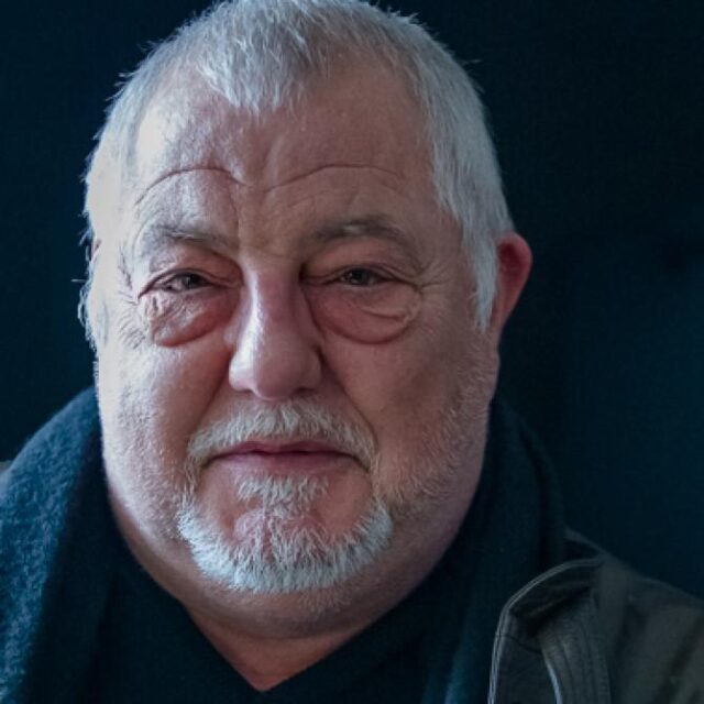 Close-up portrait of an older man with white hair and a short beard, wearing a dark scarf and jacket