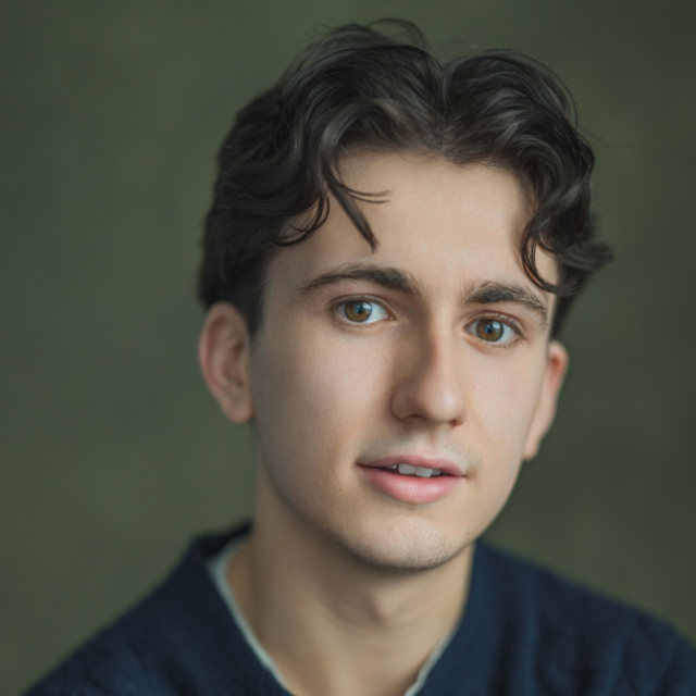 Headshot portrait of a young man with curly dark hair, wearing a navy jumper, against a green background
