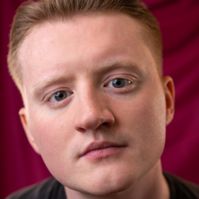 Close-up portrait of a young man with short reddish-blond hair and blue eyes, against a dark red curtain background