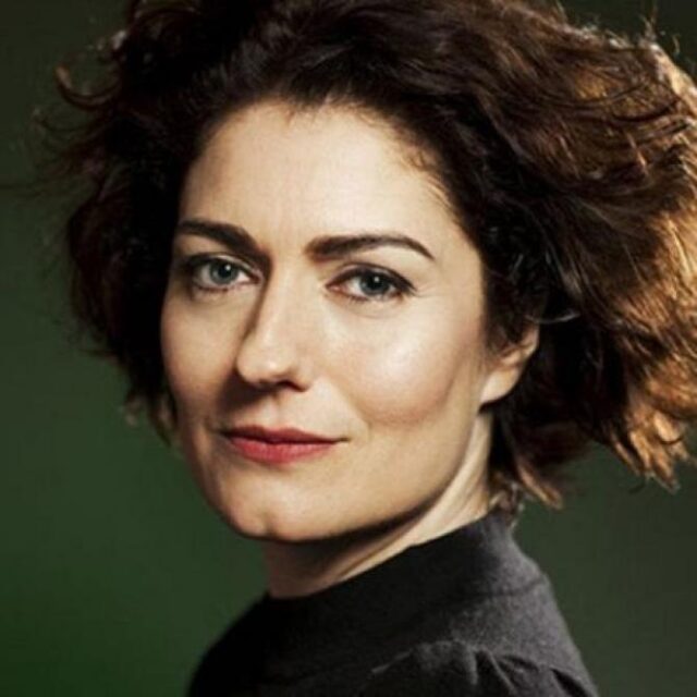 Woman with curly brown hair looking over her shoulder, wearing a black top against a green background