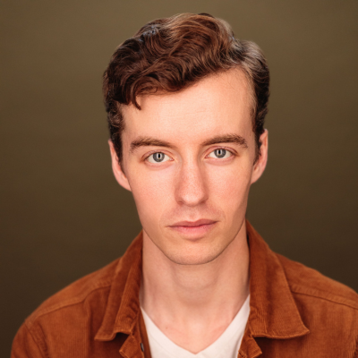 Studio headshot of a young man with wavy brown hair, wearing a rust-coloured jacket over a white top