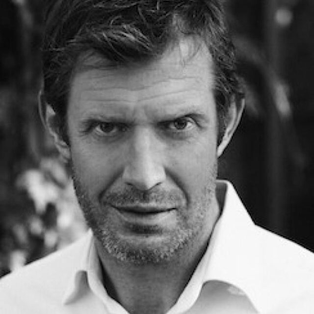 Black and white portrait of a man with tousled hair and stubble, wearing a white shirt, looking directly at the camera