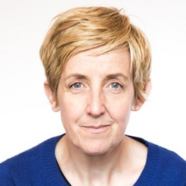 Portrait of a woman with short blonde hair, wearing a blue jumper, against a plain background