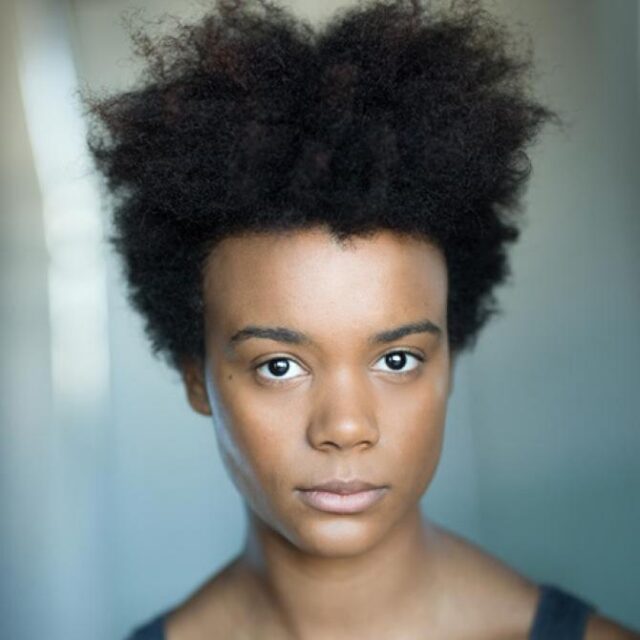 Close-up portrait of a young woman with an afro, looking directly at the camera with a neutral expression