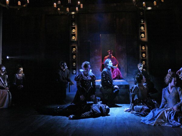 Theatre scene with actors in period costume kneeling before a robed figure on a dais, flanked by shelves of skulls under candlelit chandeliers