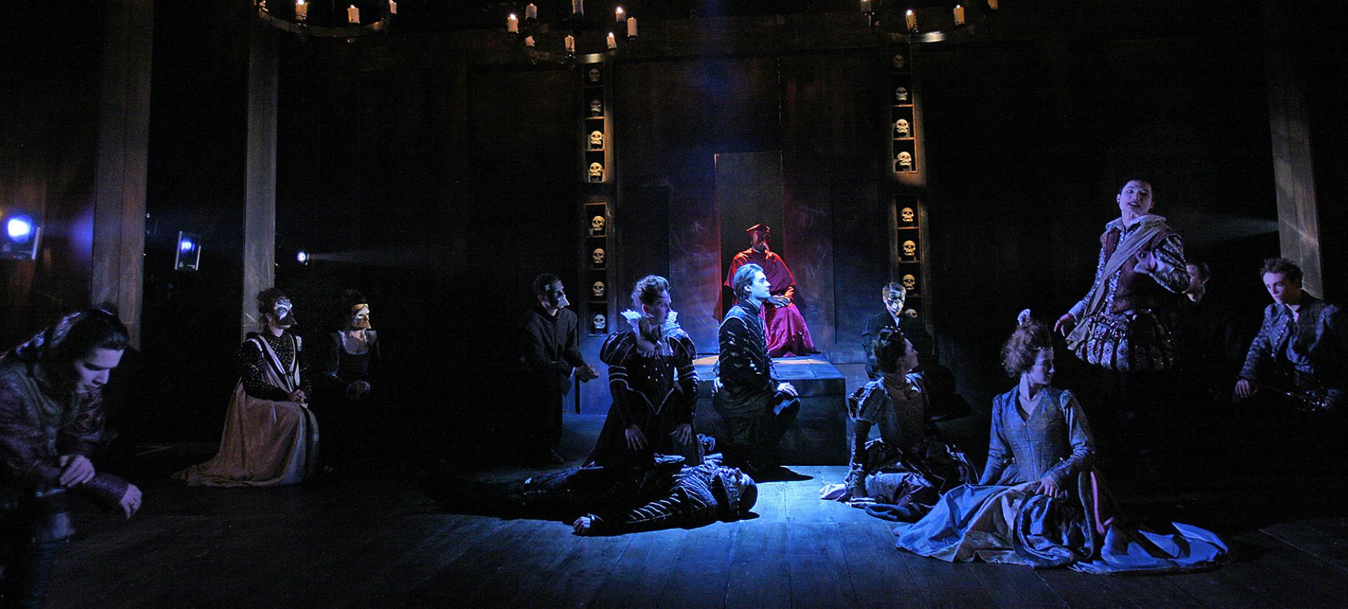 Theatre scene with actors in period costume kneeling before a robed figure on a dais, flanked by shelves of skulls under candlelit chandeliers