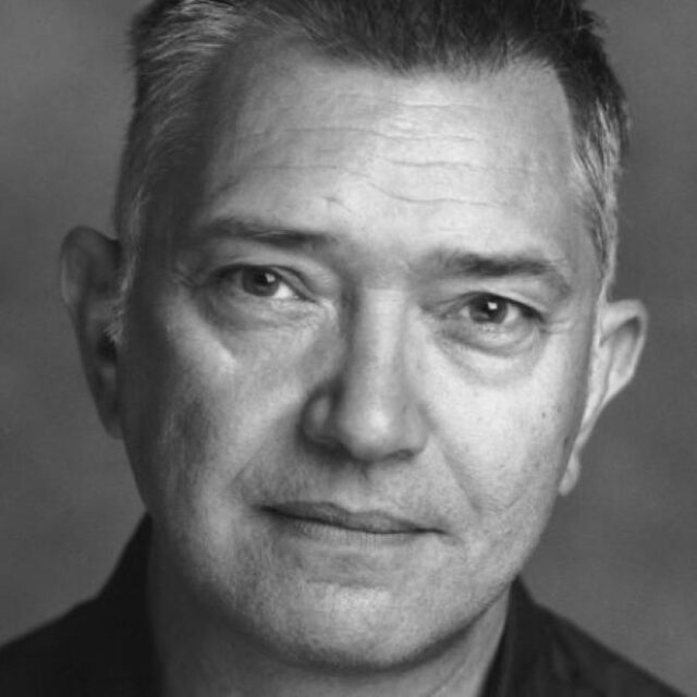 Black and white close-up portrait of a man with short greying hair, looking directly at the camera