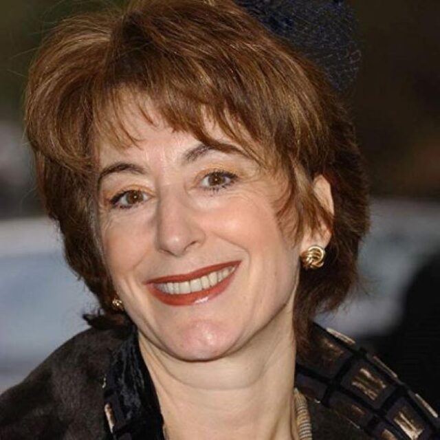 Close-up of a woman with reddish-brown hair, smiling, wearing a dark hat and patterned jacket