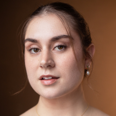 Close-up portrait of a young woman with dark hair pulled back, wearing pearl earrings, against a warm brown background