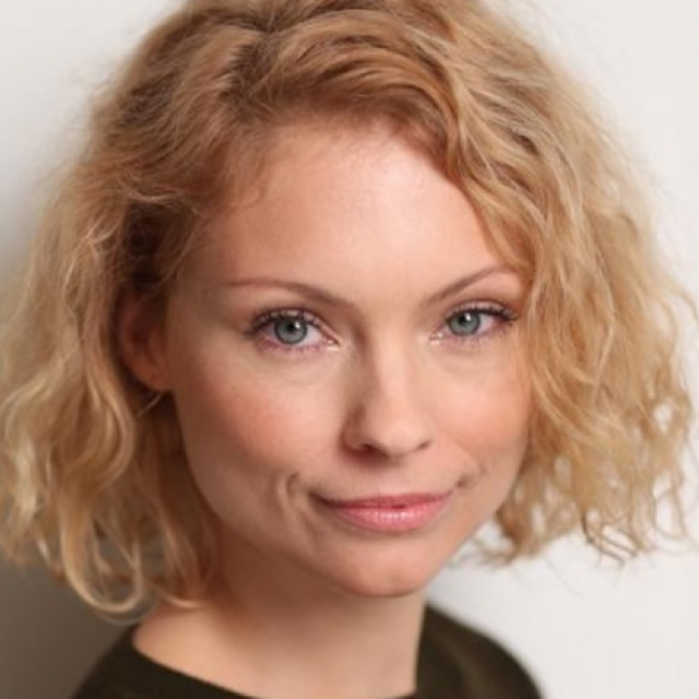 Headshot of a woman with curly blonde hair, blue eyes and a slight smile, wearing a dark top