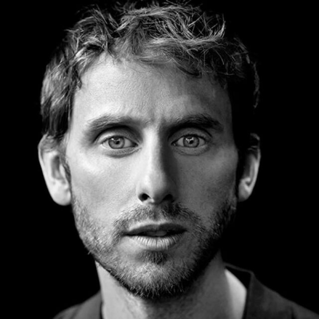 Black and white close-up portrait of a man with curly hair and light stubble looking directly at the camera