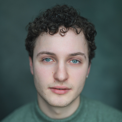 Young man with curly dark hair and blue eyes, wearing a green jumper, against a dark teal background