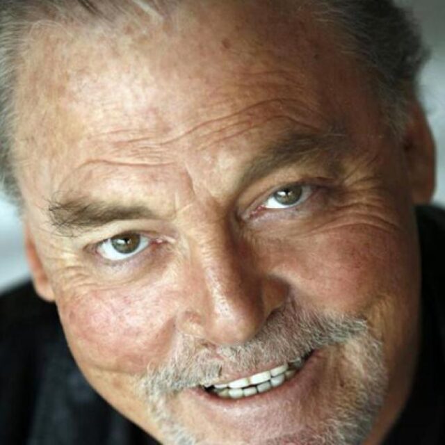 Close-up portrait of an older man with grey hair and a moustache, smiling at the camera