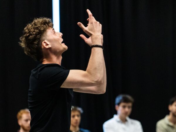 Performer with curly hair gestures dramatically upwards mid-performance, watched by an audience