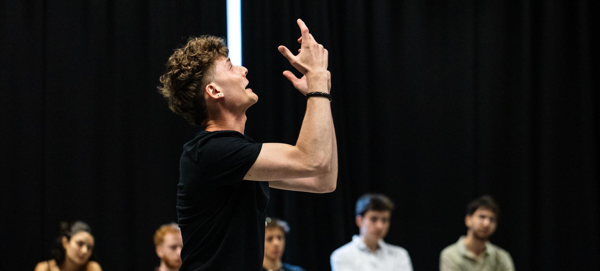 Performer with curly hair gestures dramatically upwards mid-performance, watched by an audience