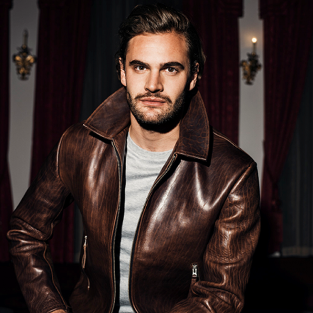 Man with tousled brown hair and stubble wearing a brown leather jacket, standing before dark red curtains