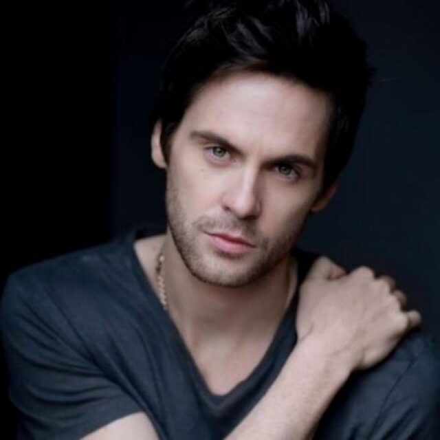 Man with dark hair and a chain necklace, wearing a dark T-shirt, looking at the camera against a dark background