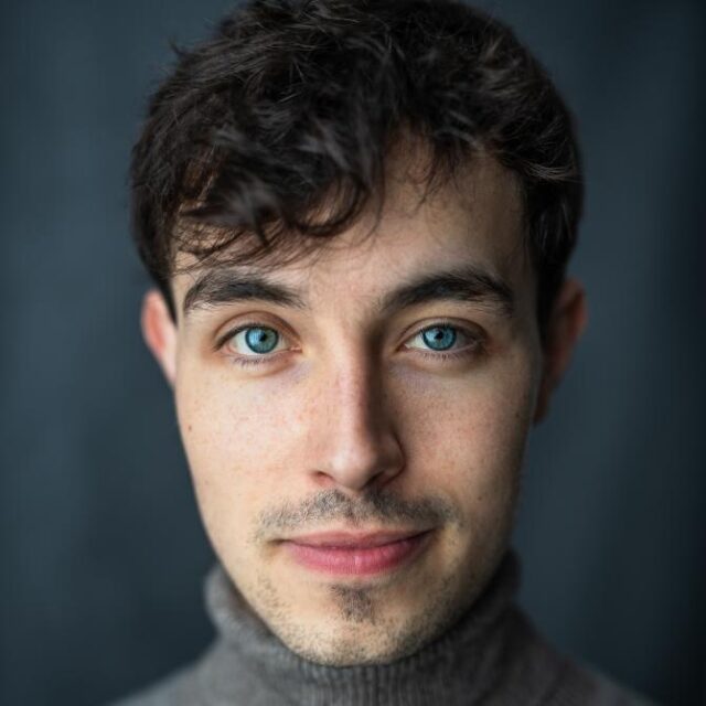 Close-up portrait of a young man with curly dark hair, blue eyes and light stubble, wearing a grey jumper
