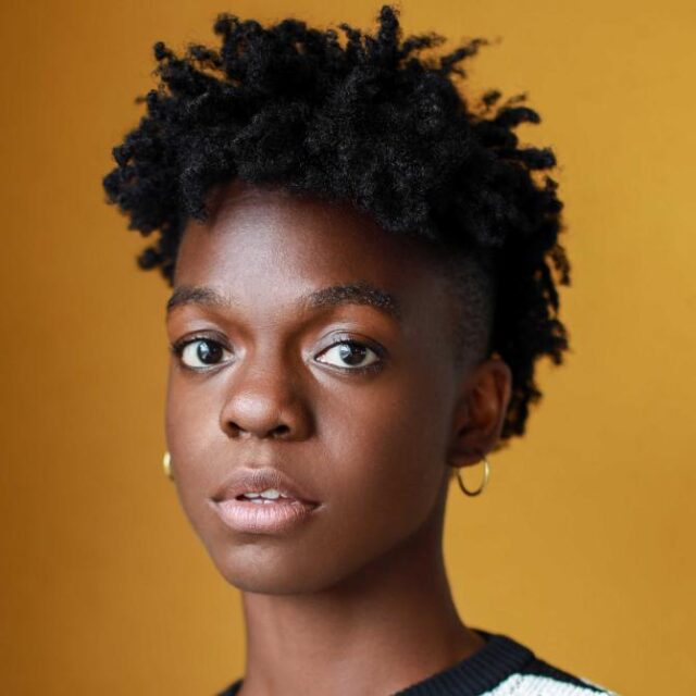 Close-up portrait of a woman with short curly hair and gold hoop earrings, against a mustard background